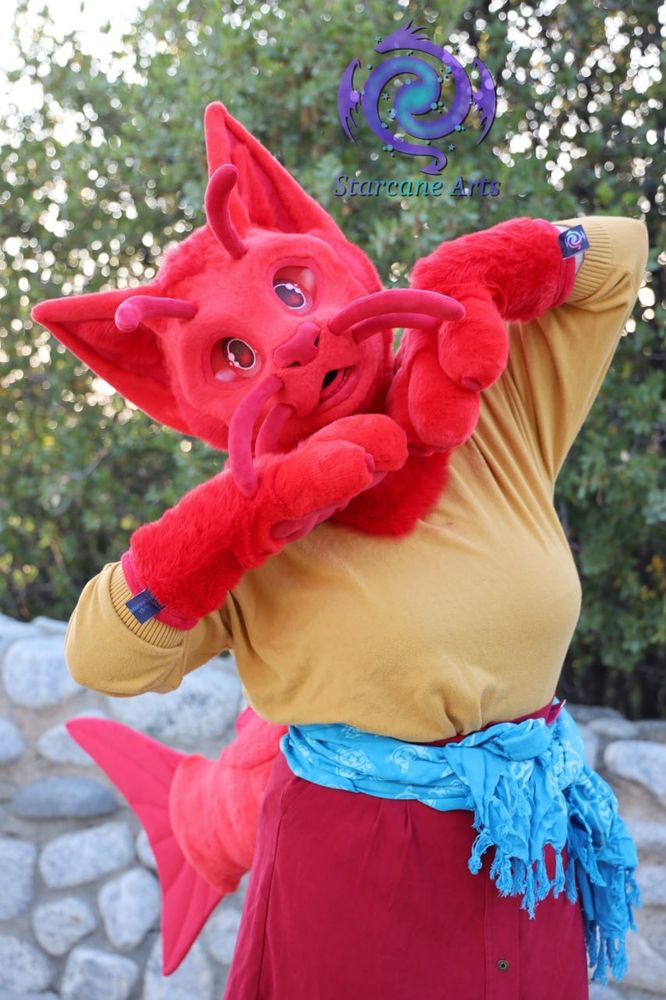 An all red "cat-fish" partial fursuiter who is posing for the camera. They are wearing a yellow shirt and a blue scarf around their waist.