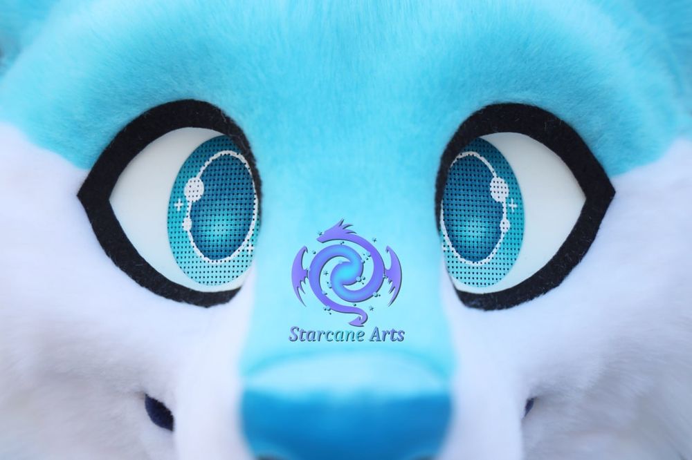 A close-up of a bright blue and white fursuit face with the blue eyes being the primary focus. A blue nose can be seen just at the bottom of the frame.

The Starcane Arts logo is visible as a watermark just above the nose. 