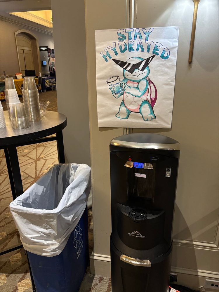 A water cooler that has a poster sized drawing of the Pokémon Squirtle (a blue turtle with a red shell) wearing sunglasses and holding a glass of water. Words above the Squirtle say “Stay hydrated”.