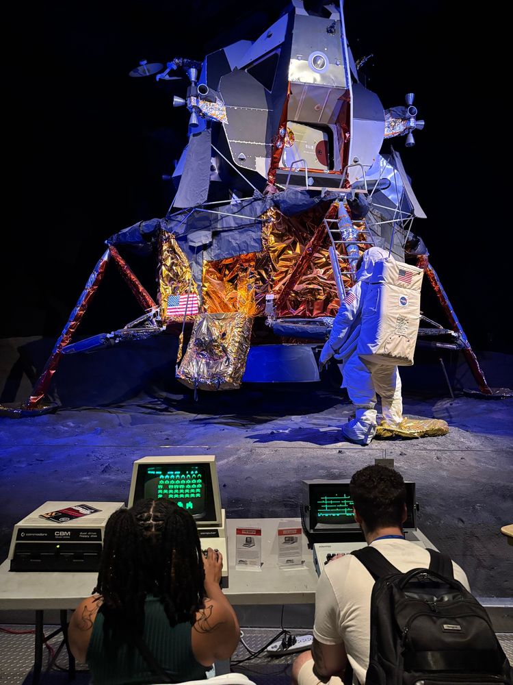 Two people are playing computer games on a desk that is directly in front of a moon lander. One of the computer games is Space Invaders (a green monitor showing rows of aliens in the sky as they march toward the player on the bottom). The moon lander is part of the aviation museum display. It has an astronaut suit that is facing toward the lander, made to look like it is approaching the nearby ladder to climb inside.