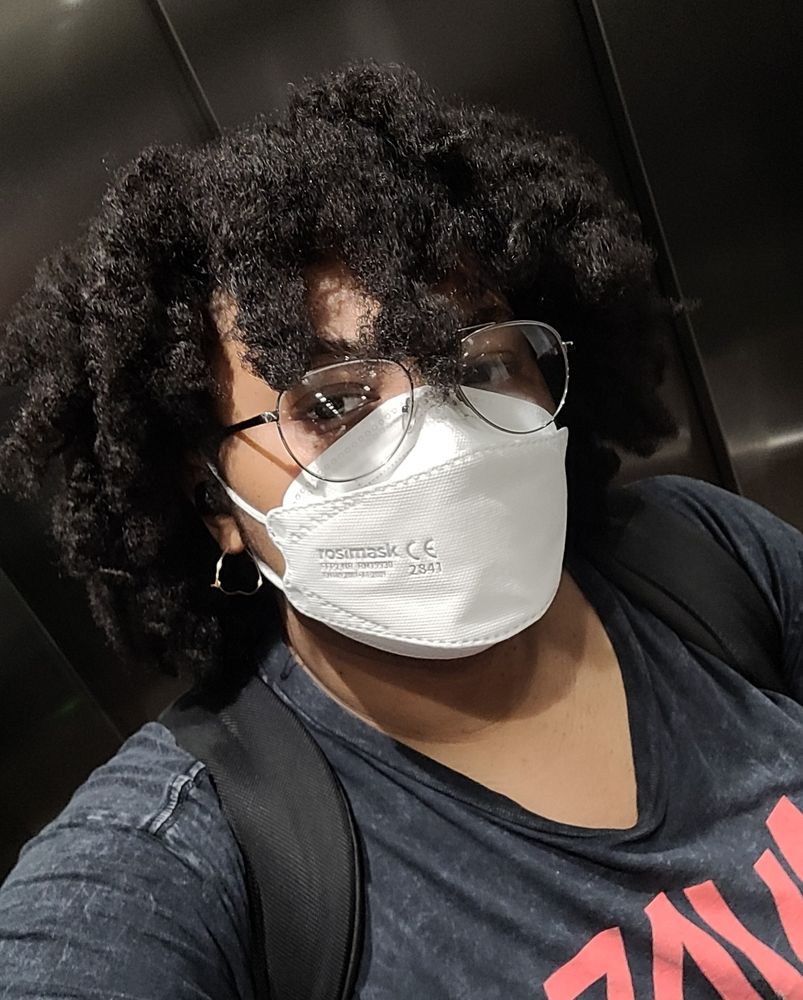 A selfie of Antoinette cut off at the bust. They have coily hair hanging around their face. They are wearing glasses, a mask and a gold hoop earring shaped like a heart. The shirt their wearing has lettering thats cut off. 
