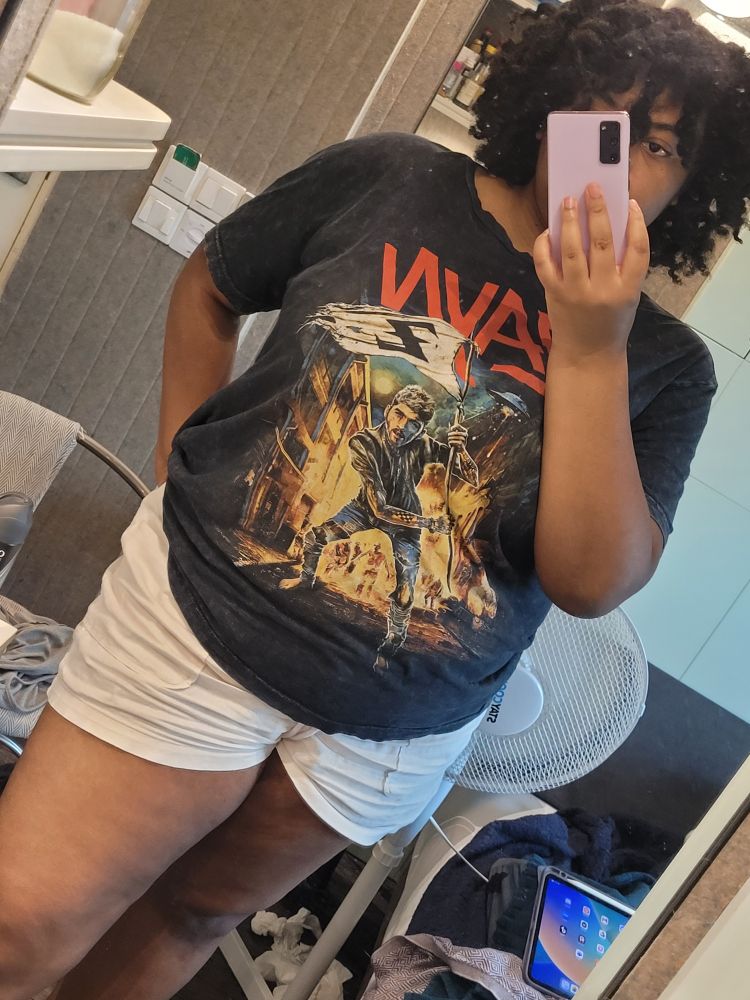 Antoinette is taking a mirror selfie in the picture. The phone covers most of their face. Their hair is coily and frames their face. They are wearing a Zayn shitt and shorts