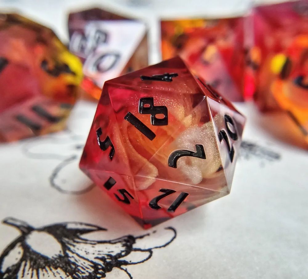 A photo of handmade D20 Sitting on a vintage botanical illustration, it has a green leaf nestled next to the die.  The dice are a bright red to amber gradient that is dotted with red micro glitter and has swirls of white smoke inside of them. They are inked in a gold to black shifting ink.