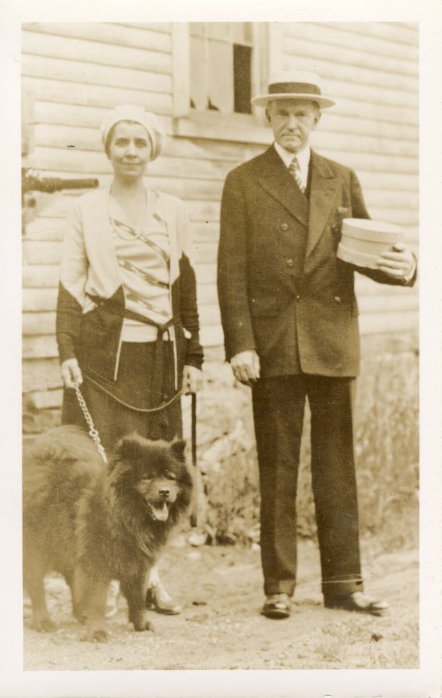 Tiny Tim, a brown chow dog is standing and on a leash held by Grace Coolidge. She is standing in front of the side of a  house with clapboard siding and a window and wearing a light color top and dark shirt and white hat.  Standing next to her is Calvin Coolidge in a dark color suit and straw hat.  He is holding a round wood box, probably a cheese box. 