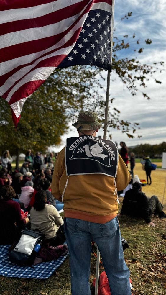 A man wearing a Veterans for Peace symbol holding an upside down American flag while a speaker addresses a large crowd.