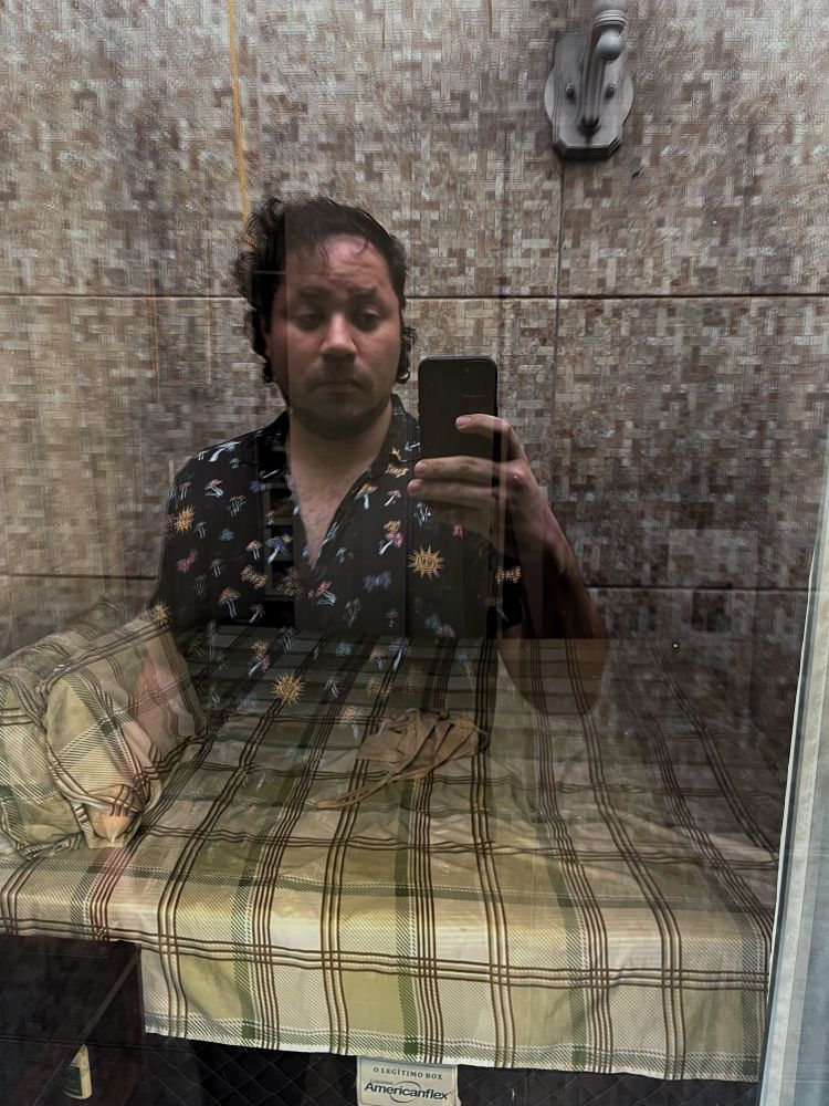 Selfie num vidro espelhado, no plano da frente tem eu, uma pessoa branca, usando uma camisa azul com várias estampas coloridas meio aberta, segurando um celular. Na parte de trás do reflexo, uma cama. 