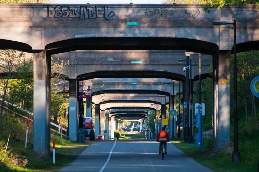 The Midtown Greenway, a below-grade pedestrian and bike path in Minneapolis. The path goes below the cross streets. This shot shows many bridges from path level that makes it look like a tunnel, although there is open sky between each bridge. Each side of the path is flanked by green space. A biker is heading away from the camera. There is grafitti on the nearest bridge. 