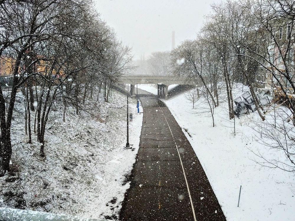 A snowy shot of the Midtown Greenway in Minneapolis, a bike and pedestrian path. Snow is falling and blankets the green spaces that flank the path. The shot is taken from above on a bridge, and another bridge is visible in the background as the path curves to the left.