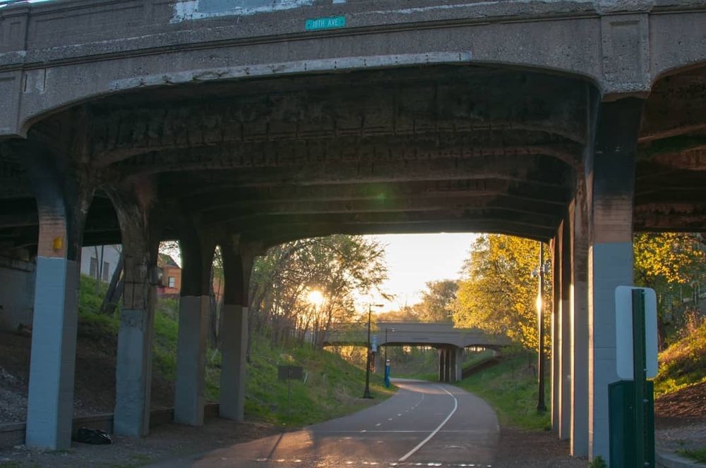 A shot on the Midtown Greenway in Minneapolis. It is a pedestrian and bike path. The shot is from path level with two bridges going over it as it curves to the left. The sun is just rising and shining through trees in the green space that flanks both sides of the path. 