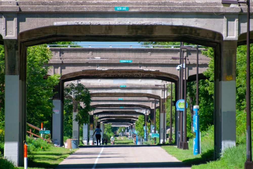 The Midtown Greenway, a below-grade pedestrian and bike path in Minneapolis. The path goes below the cross streets. This shot shows many bridges from path level that makes it look like a tunnel, although there is open sky between each bridge. Each side of the path is flanked by green space. A runner is headed away from the camera. 