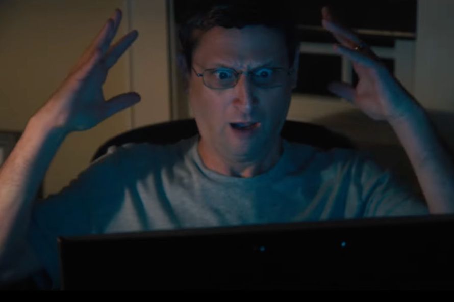 Tim Robinson sits at a computer monitor with his hands in the air with a shocked look on his face.