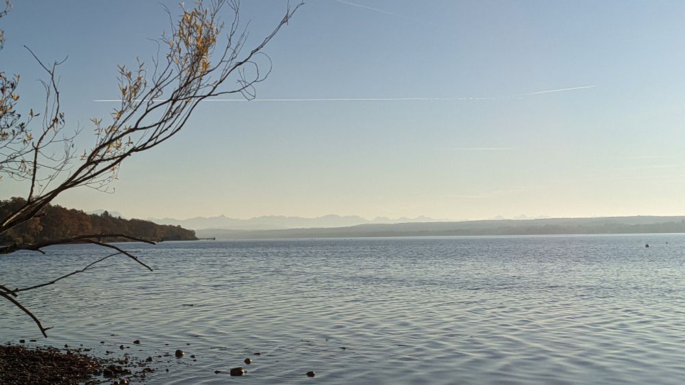 Landschaftsbild: Blick über den Ammersee. Am Horizont sind die Alpen zu sehen, darüber wolkenloser Himmel. 