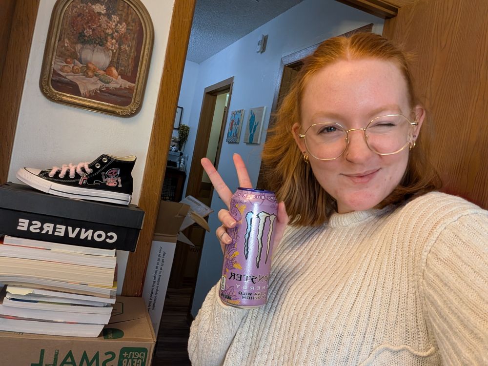 me, holding a monster and peace-signing. red hair, basically non-existent eyebrows, glasses, and blotchy skin lmao.