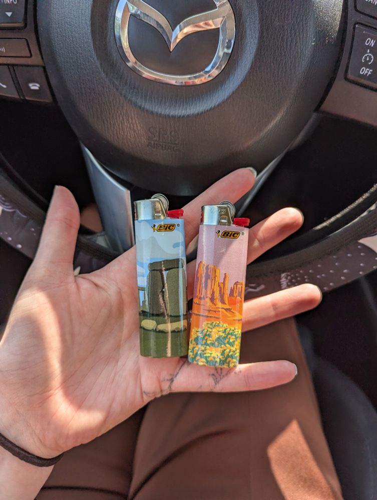 two bic lighters in my hand with my steering wheel behind. the lighter on the left has Stonehenge on it. the lighter on the right has Monument Valley on it.