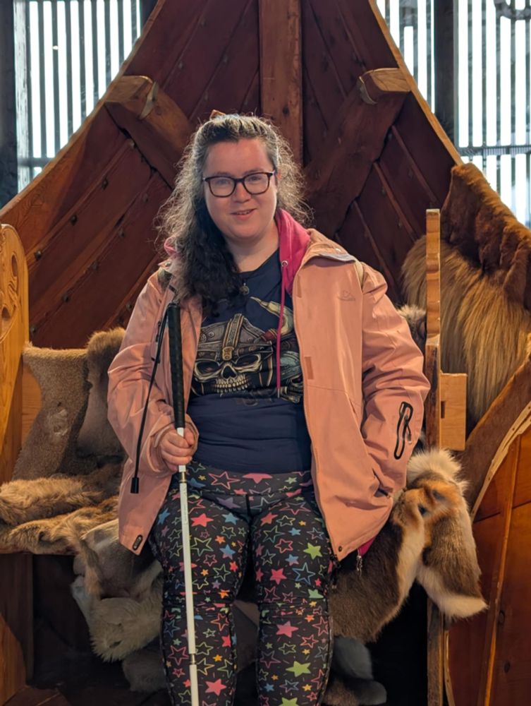 Me sitting on a viking ship in the captions chair wearing a t-shirt with s viking on and a pink raincoat. Smiling at the camera holding my long cane.