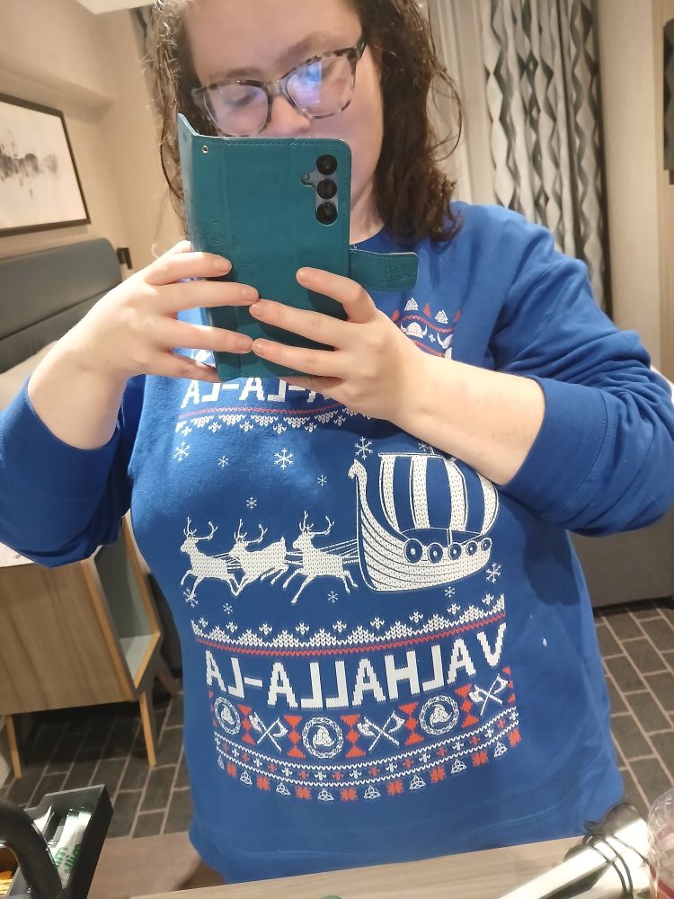 A picture of me smiling at the camera wearing a dark blue norse Christmas jumper. It says fa la la la valhal la la around an image of a viking skip being pulled by reindeer
