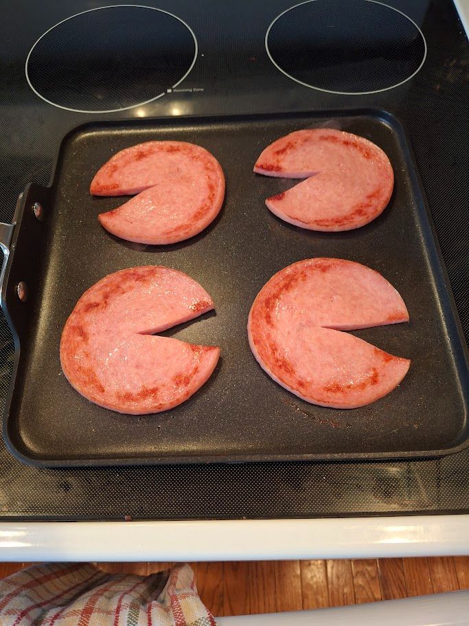Four slices of pork roll frying. They have slits that make them look like Pacman.