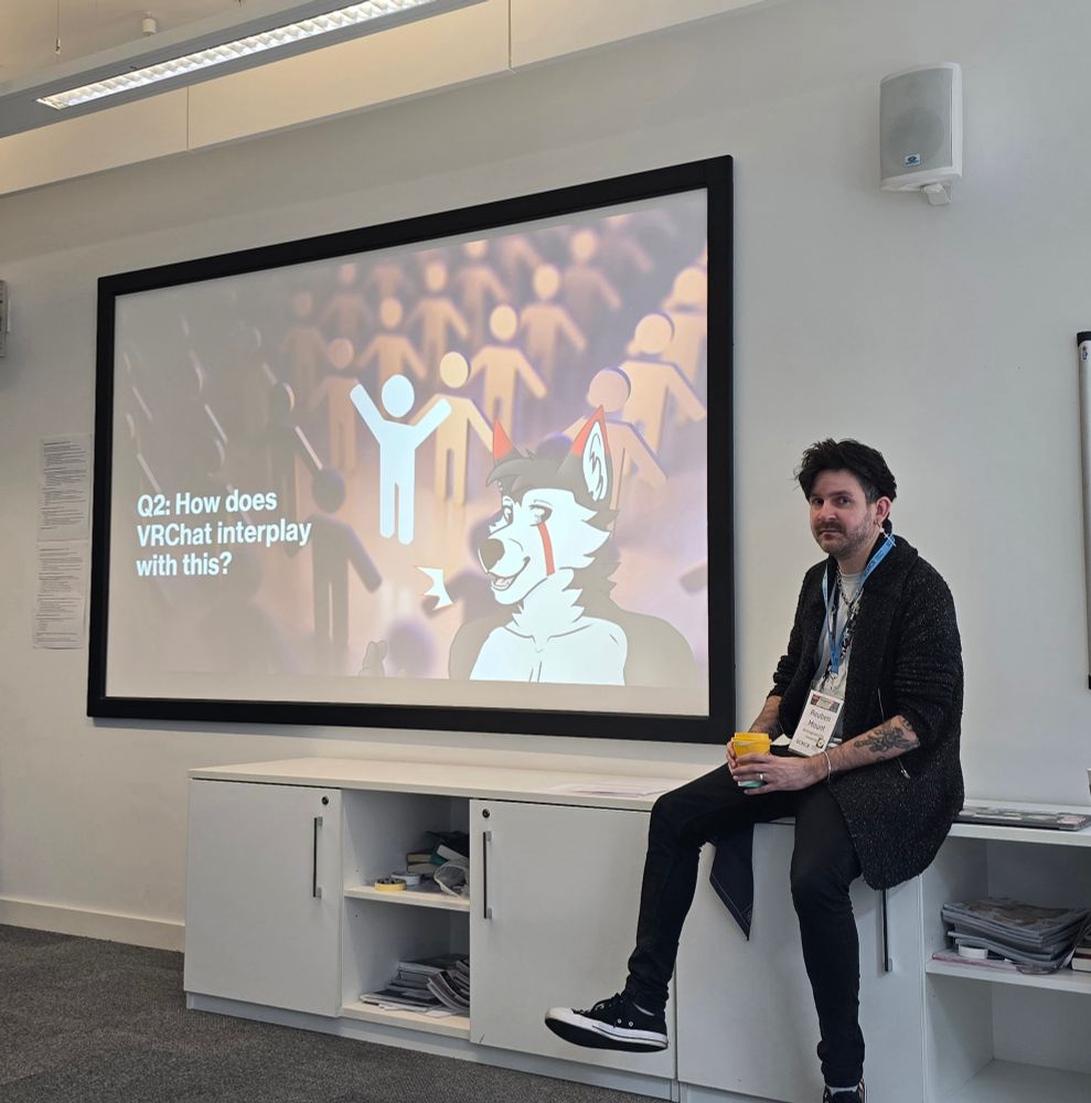 Reuben Mount is sitting on the edge of a white cabinet in a classroom or conference room. He is holding a cup and wearing a lanyard with a name badge. Behind him, a large screen displays a slide that reads "Q2: How does VRChat interplay with this?" alongside a cartoon drawing of a humanoid husky character - his fursona, Vanguard. The background of the slide features stylized figures of people in muted colors. The room has a modern design with a whiteboard and papers on the wall.