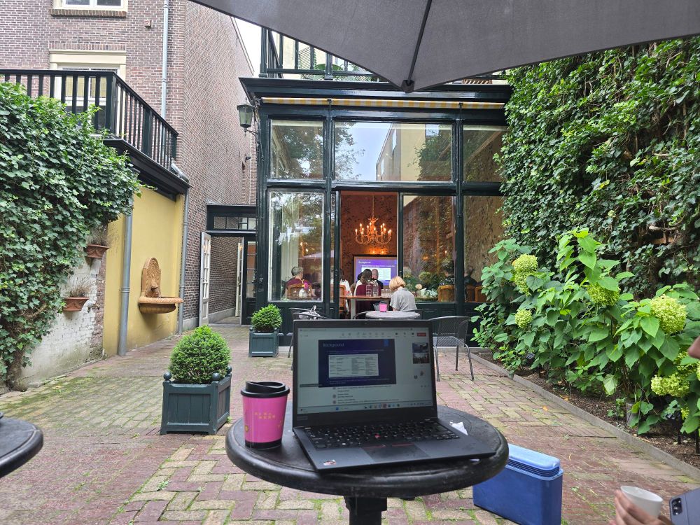 Outdoor courtyard seating area with brick flooring, green plants along the wall, and a laptop on a round table under a large umbrella. The laptop screen shows a presentation slide. In the background, through large glass doors, people are sitting indoors at a table beneath a chandelier, and are watching the same presentation slides projected to a full room of people. A decorative wall fountain and potted shrubs add detail to the courtyard.