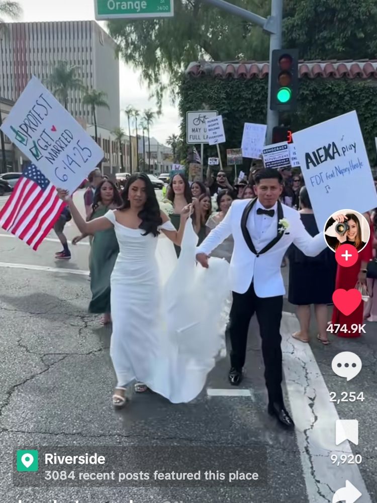 Tiktok screenshot of a bride and groom, both Mexican-American, in a gown and tux on the streets of Riverside holding signs that say "protest, get married" and "Alexa, play FDT"