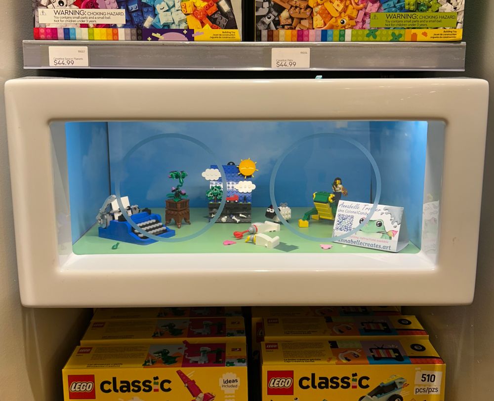 Various LEGO creations in a display case. Includes a road trip scene, a frog, a mini typewriter, paint tubes, a baby sheep and a minifigure representing the artist.