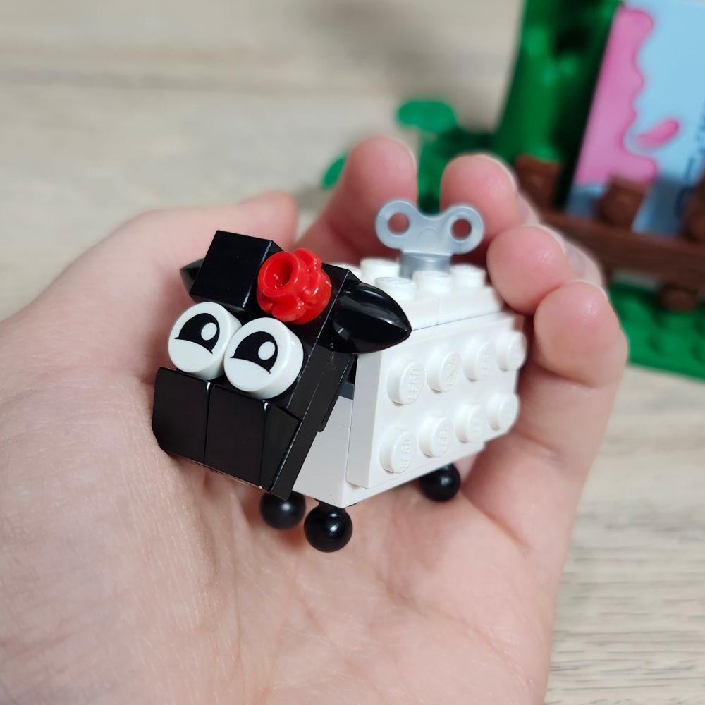 Little LEGO sheep being held in the palm of an hand. It has a red flower on the head and a toy winder key on the back.