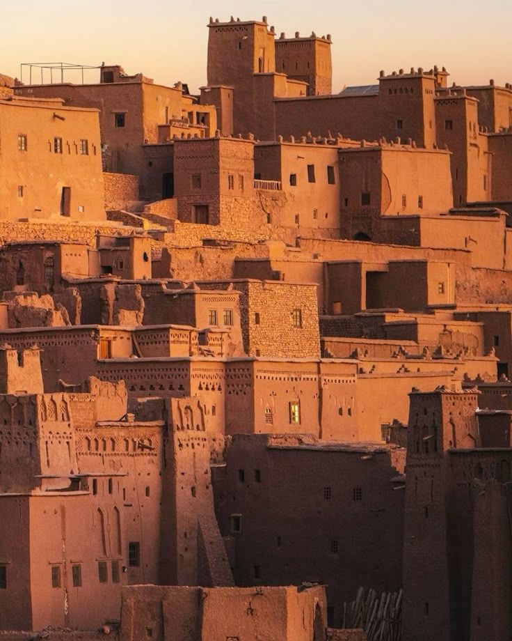 A scenic shot of Morocco's square, carved buildings catching the golden rays of a setting sun. They're of different heights and widths. With the different elevations, you get to see them stack upon each other like the sea. At s closer look, you get to see their walls carved designs near the top of their flat roofs. A strip of pale blue and yellow sky lies at the top of the photo.