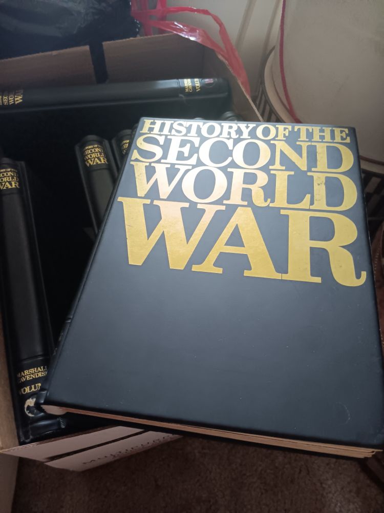 A black binder with bold letters "History of the Second World War" is displayed upon it. It lies diagonally on a box of the other volumes of the same binding. The box is nestled with other packed away items. The carpet is beige.