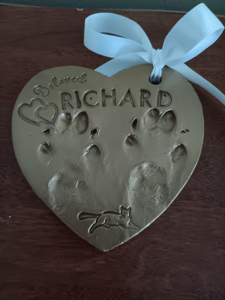 A golden paw print impression of Richard's two paws shaped like a heart. Scratched in it was his name, with two stylized hearts, 'beloved', and an image of a cat lounging at the bottom. There's a white ribbon tied at the corner.