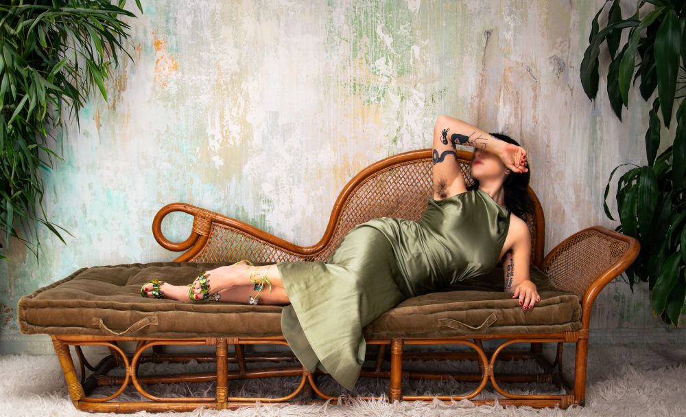 A curvy, tattooed brunette woman lounges on a velvet daybed with a rattan backing. She wears a green silk dress and green sandal heels. Plants line the sides of the frame and the background is green, gold, and white. One arm covers her face, leaving her nose and lips visible