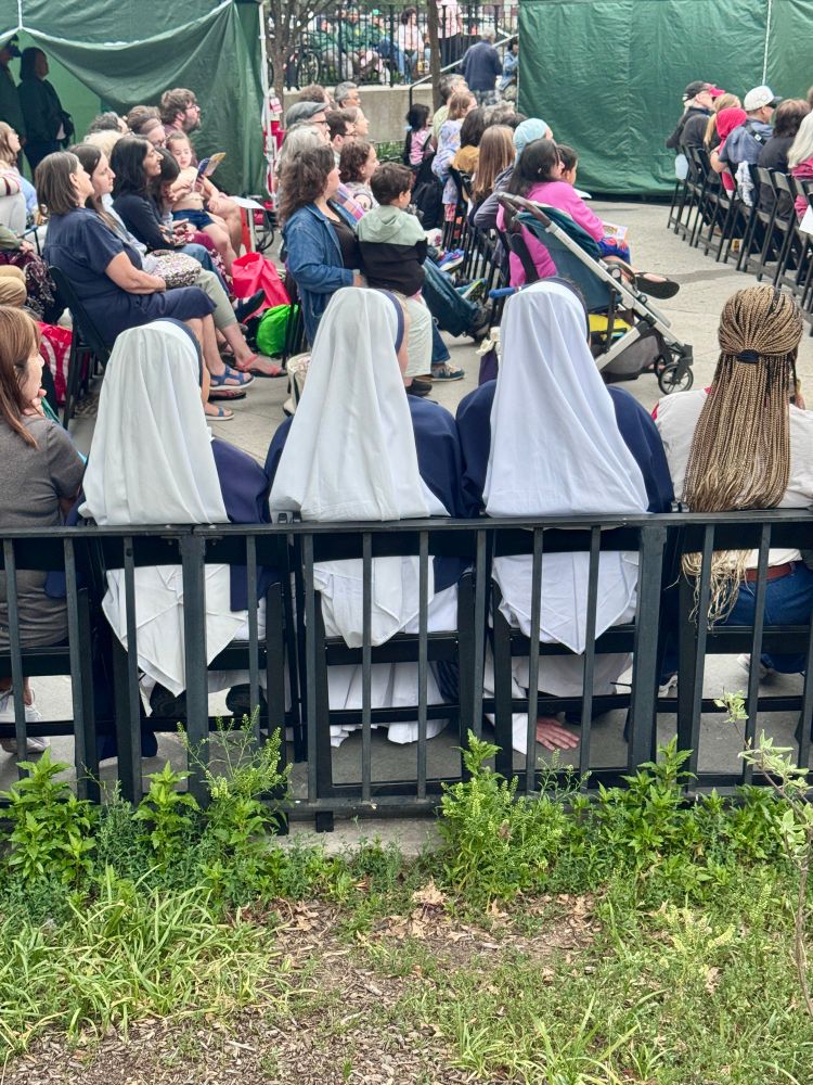 Three nuns in blue and white habits with a hand visible under one nun’s chair. 