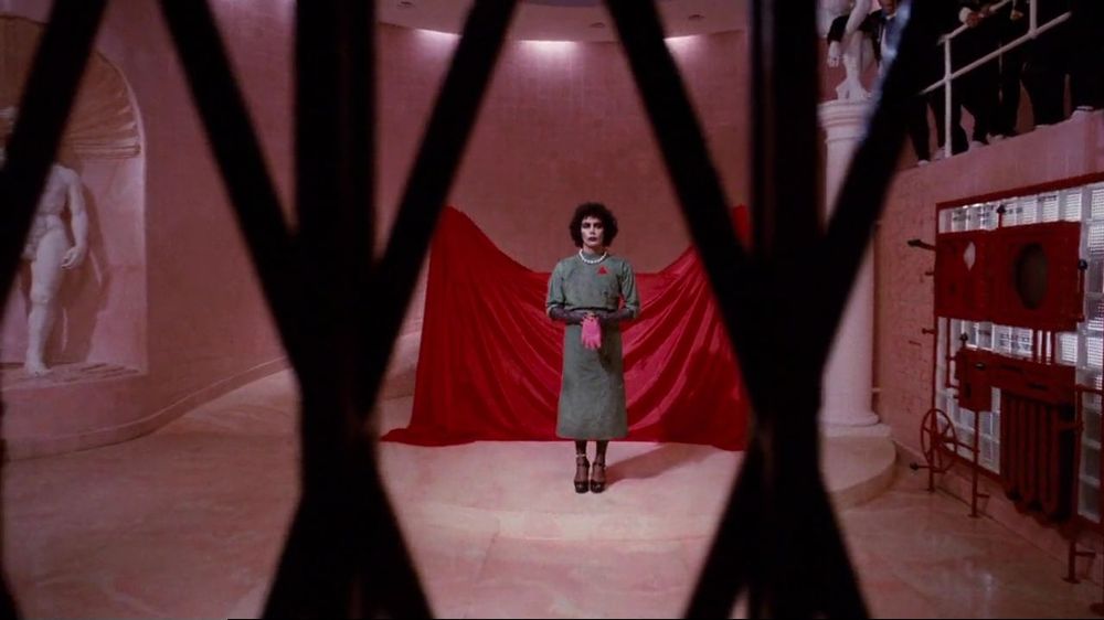 Visão de Dr. Frank-N-Furter (Tim Curry) por entre as grandes de proteção de um elevador. Frank se posiciona ao centro da cena. Está em uma sala toda rosa e com um pano vermelho que cobre algo logo atrás de sua figura. À sua esquerda, uma estátua de mármore. À direita, um painel de controles e alavancas, todas vermelhas. Logo acima, uma plateia de pessoas em trajes sociais.