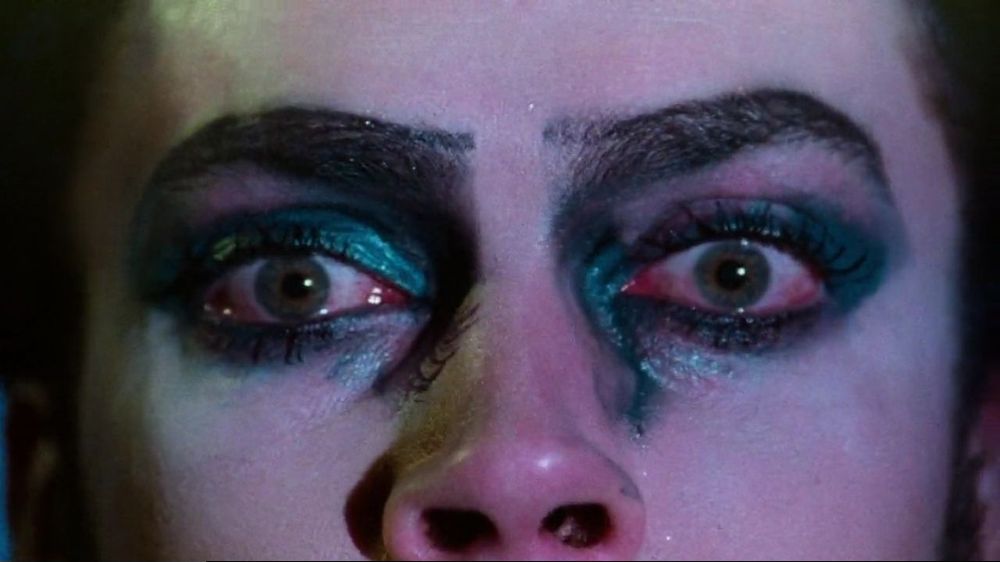 Plano detalhes dos olhos de Frank-N-Furter (Tim Curry) que estão vermelhos e orçados por uma maquiagem azul cintilante. Frank traz medo e consternação no olhar.