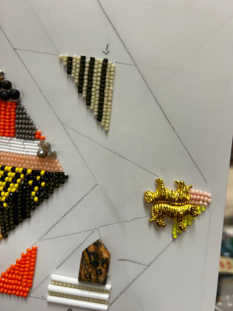 A third crow of a complex beading project, about 4x4 inches, in two stacked chevrons, with colors of gold, orange, grey, orange-pink and white. Highlight? Two gold tigers near the center-tip. 