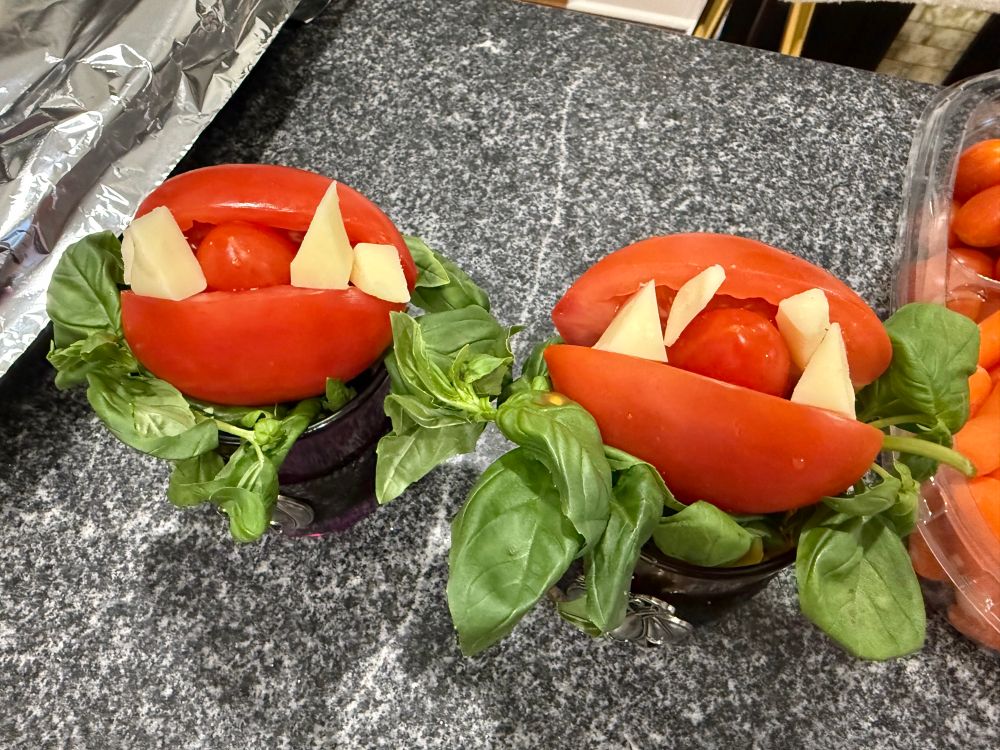A roma tomato that has been sliced like a hotdog bun and placed in a cup on a bed of basil. Inside the cut there is a cherry tomato that forms the tongue of the venus fly trap, then pieces of cheese have been cut into fangs and placed into the tomato to line the mouth.