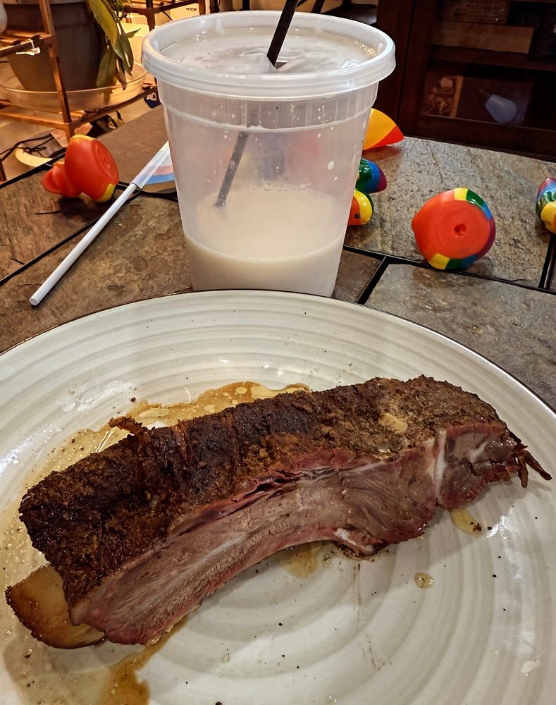 A dinner plate with one massive BBQ beef rib on it. Behind the plate is a 32 ounce deli cup of horchata that has been 2/3 consumed. There is a pride flag and some rainbow rubber ducks scattered on the table.