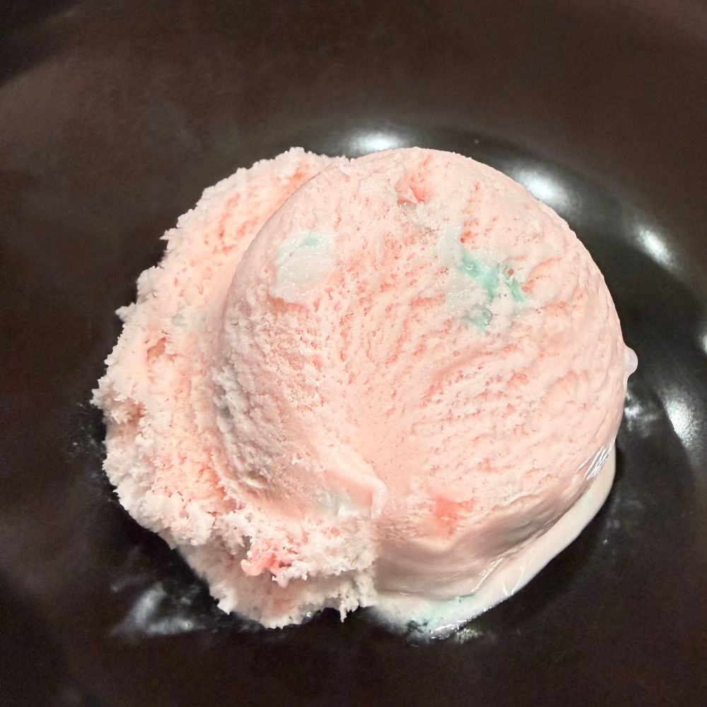A single scoop of peppermint stick ice cream in a bowl. It's a light pink ice cream with bits of red and green peppermint candy in it.