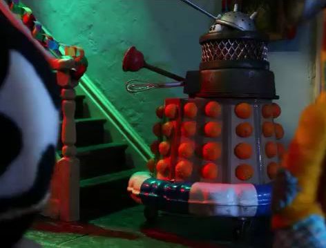 An image from the Shaun the Sheep episode Party Animals. One of the sheep has come to a fancy dress party as a Dalek, with a costume made out of bits of junk. Only the eyes of the sheep can be seen under a colander 'head'. The Dalek is trying, unsuccessfully, to go up some stairs.