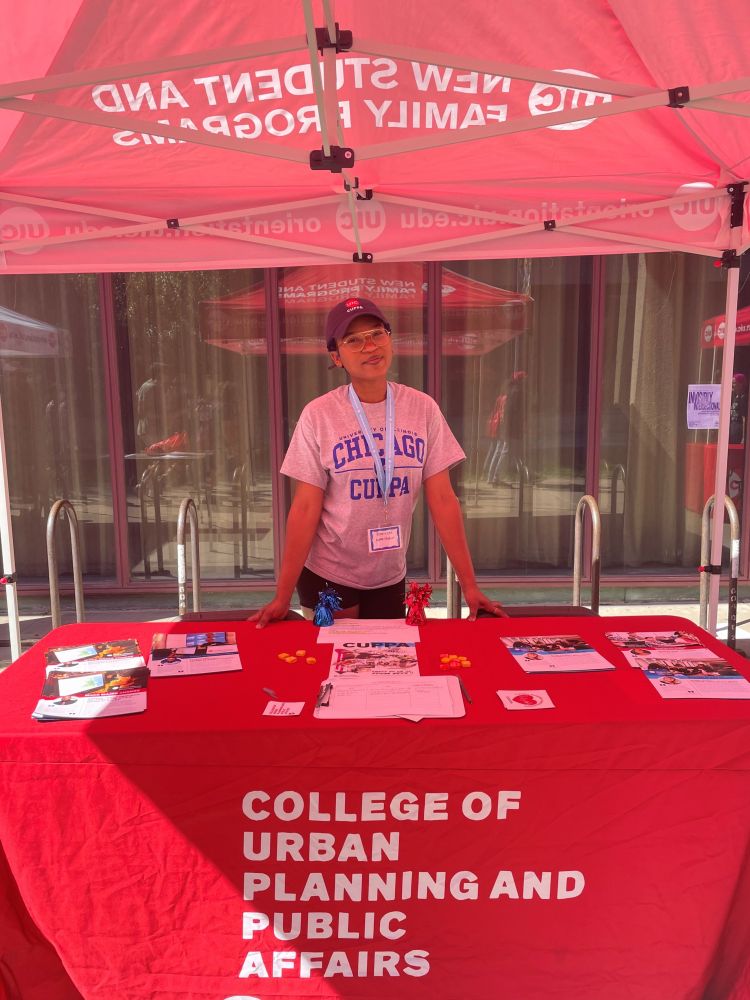UIC CUPPA information booth with a red table cloth and a student representative providing details on public policy, public administration, urban planning, civic analytics, and city design graduate degrees and public policy and urban studies undergraduate degrees from UIC.