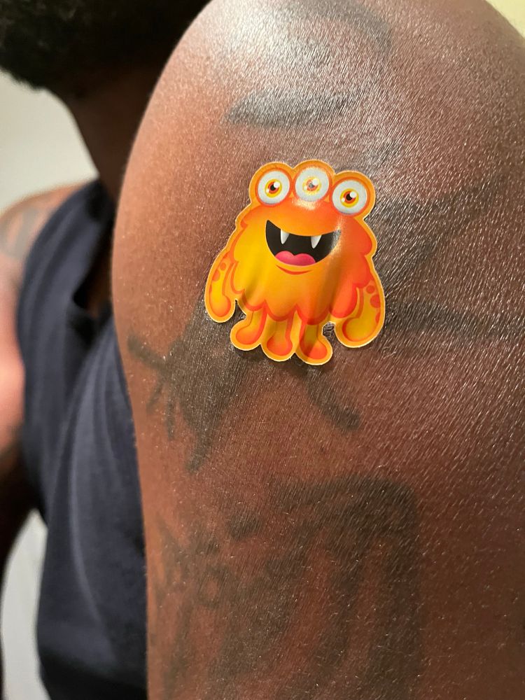 A picture of an orange monster bandage on my arm. The monster is smiling and has three eyes, three legs, and two arms.