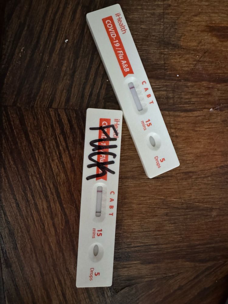 Two Covid tests on a table, both test for two types of flu, and Covid-19. One is negative for all, and one is positive for Covid and labeled FUCK in all caps with a sharpie