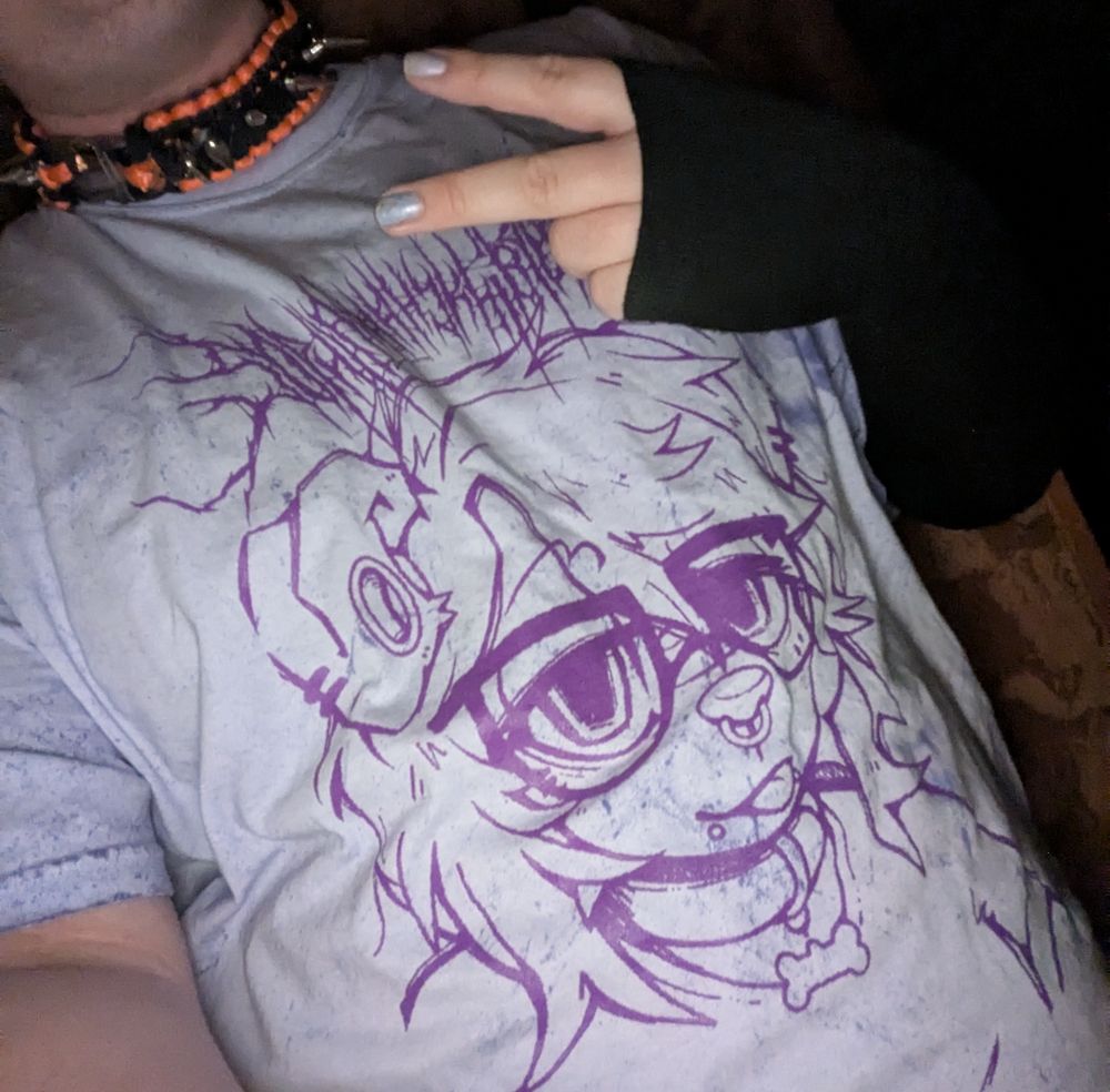 a white person is wearing a purple tshirt, the shirt depicts an anthropomorphic dog wearing glasses. the person wearing the shirt is wearing a spiked black and orange collar and a  black fingerless glove on their left hand, which is giving a backwards peace hand sign. you can see their nail polish matches the shirts' shade of purple.