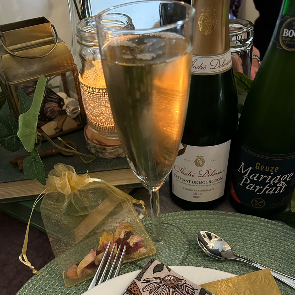 Picture of a glass of bubbly surrounded by:
- Top left: A stack of old books on top of which was an old key, a blonde-decorated glass with a candle in it and a faux-ivy wreath
- Top right: A small bottle of Cremant de Bourgogne and a small bottle of gueuze called "Mariage Parfait"
- Bottom: A near-empty goldynet bag of dried rose petals (to throw on the bride & groom as they did their post-ceremony walk); cutlery & plate on a green round table cover; and on top one gold and one nature-themed napkin (bc the couple is very outdoorsy)