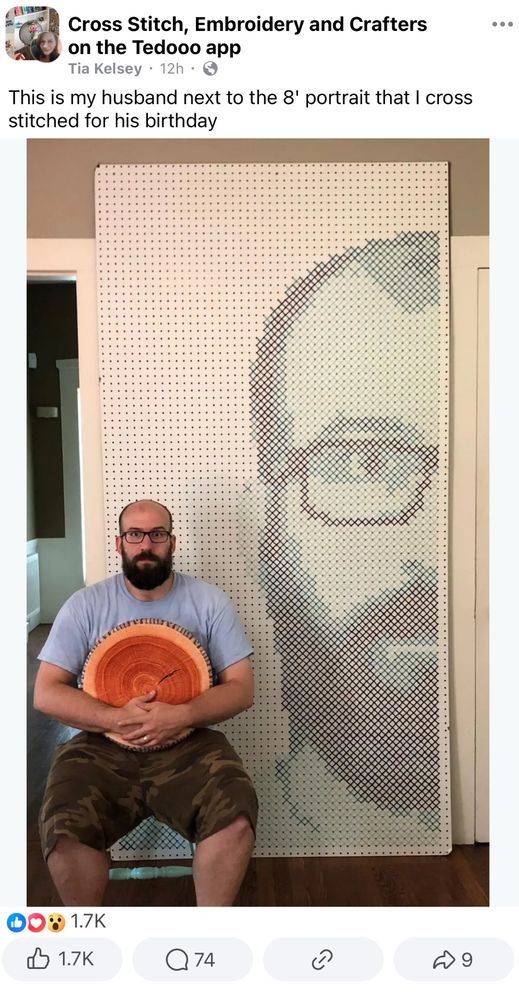 screenshot of a facebook post from Tia Kelsey in a group called Cross Stitch, Embroidery and Crafters on the Tedooo app. the post includes a photo of a bald white man sitting beside a berry large cross stitched representation of his face in blue yarn on white peg board, and a caption that reads “This is my husband next to the 8' portrait that I cross stitched for his birthday.” The post has 1.7k likes.