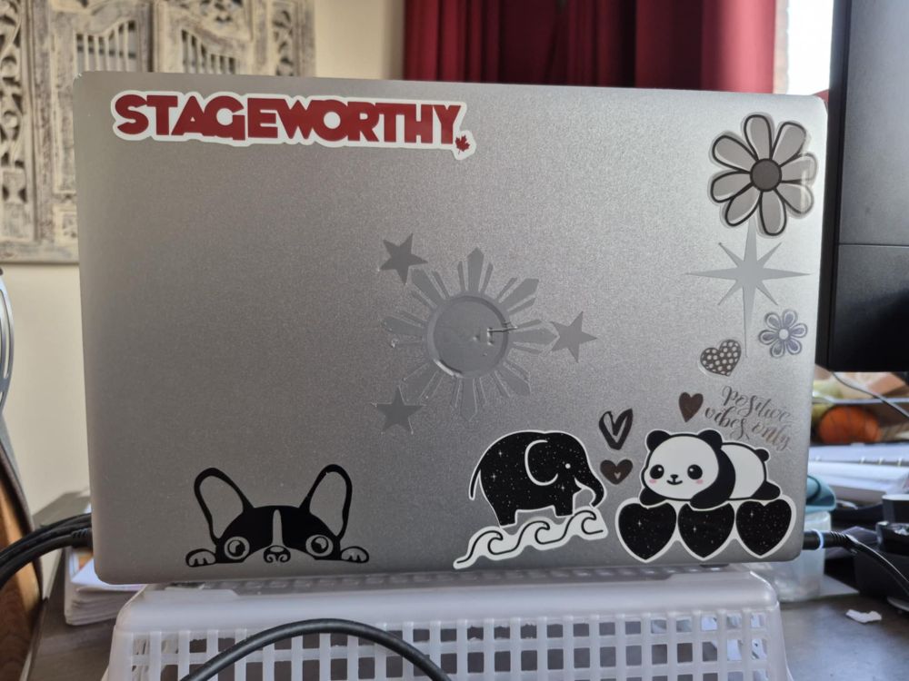 Laptop cover with stickers. Black, grey and white flowers, Philippine Sun and Starts, hearts, a peeking French Bulldog, an elephant standing on waves, a panda bear lying on hearts. A red and white logo of Stageworthy with a Canadian maple leaf.