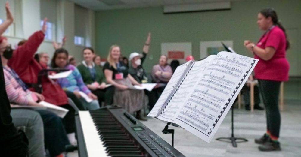 A group of people are participating in a choir practice, with some raising their hands enthusiastically. A person stands in front leading the session, near a piano and a music stand displaying sheet music.