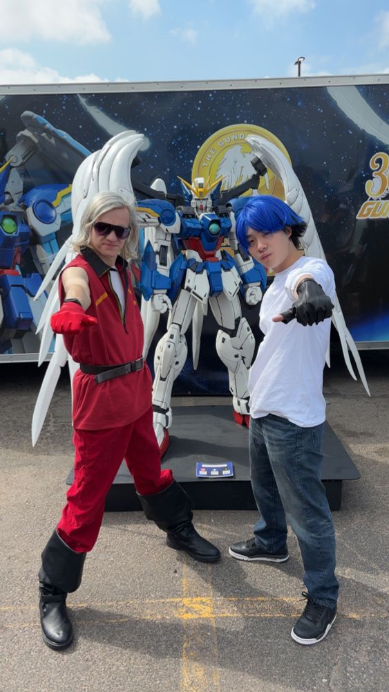 A cosplay of Quattro and Suji infront of a statue of the Gundam Wing Zero. Both cosplayers are going the "mission accepted" pose from Gundam wing