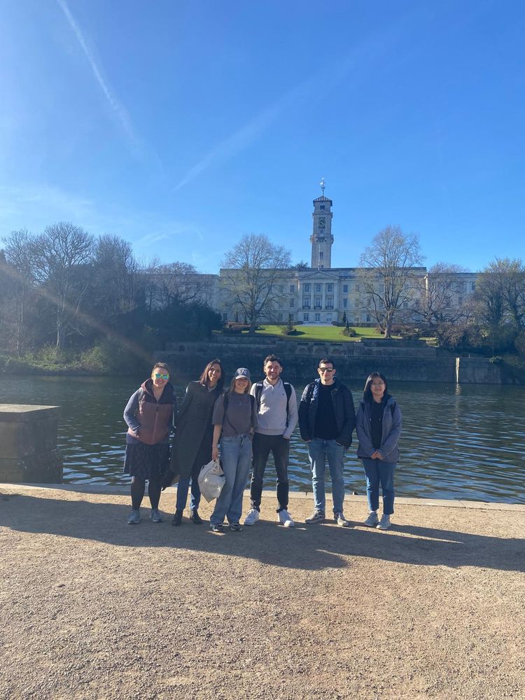 A group of people stand in the sunshine in front of a lake, the university of Nottingham Portland building can be seen in the background. 