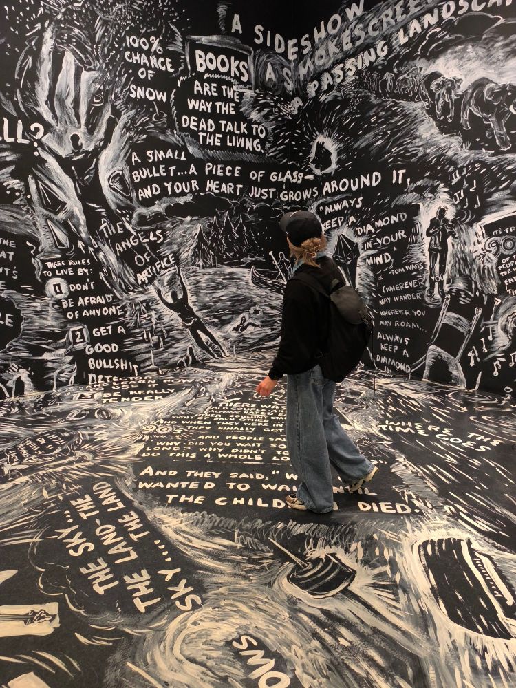 A woman stands in a room, the entire walls and floor are painted black with white text. The room is a work of art from Laurie Anderson entitled "Four Talks"