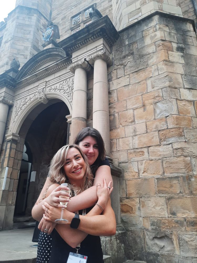 Two smiling women embrace on the steps to a castle. One has her arms around the other and holds a prosecco glass. 
